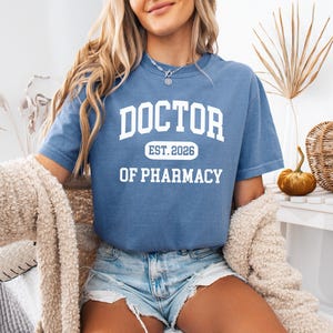 May include: A blue t-shirt with the text "DOCTOR EST. 2026 OF PHARMACY" in white block letters. The shirt is short-sleeved and has a crew neck. The person is wearing denim shorts and a cream-colored sweater.