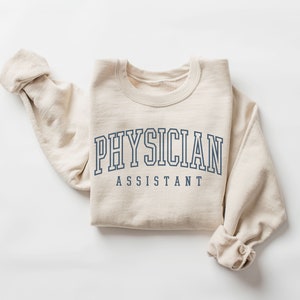 K&ouml;nnte beinhalten: Ein beiger Pullover mit dem Aufdruck "PHYSICIAN ASSISTANT" in Marineblau.