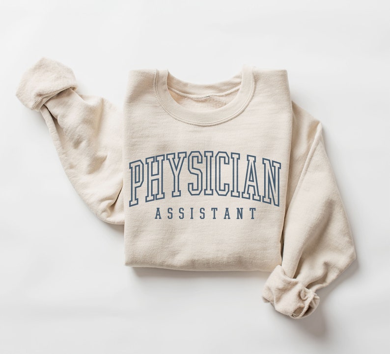 K&ouml;nnte beinhalten: Ein beiger Pullover mit dem Aufdruck "PHYSICIAN ASSISTANT" in Marineblau.