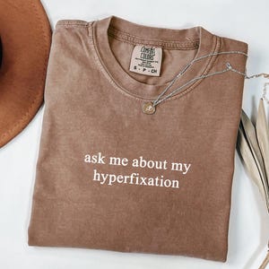 Peut inclure: T-shirt marron Comfort Colors avec le texte blanc « ask me about my hyperfixation ». Un collier argenté avec un petit pendentif est posé sur le t-shirt. Un chapeau marron et une plante séchée sont en arrière-plan.