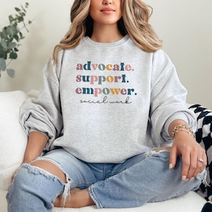May include: A light gray sweatshirt with the words "advocate. support. empower. social work" in colorful text. The sweatshirt is worn with ripped blue jeans. The overall theme is social work.