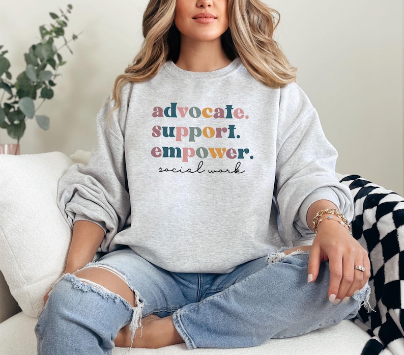 May include: A light gray sweatshirt with the words "advocate. support. empower. social work" in colorful text. The sweatshirt is worn with ripped blue jeans. The overall theme is social work.