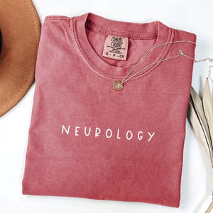 K&ouml;nnte beinhalten: Ein rotes T-Shirt mit dem Wort "NEUROLOGY" in wei&szlig; auf der Vorderseite. Das Shirt ist aus weichem, bequemem Material und hat eine lockere Passform.