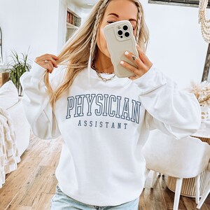 K&ouml;nnte beinhalten: Wei&szlig;er Pullover mit blauem Text, der "PHYSICIAN ASSISTANT" lautet.