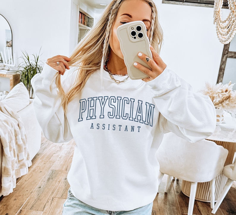 K&ouml;nnte beinhalten: Wei&szlig;er Pullover mit blauem Text, der "PHYSICIAN ASSISTANT" lautet.