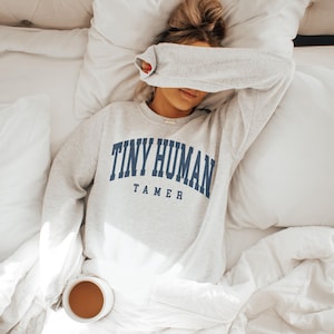 May include: A light gray sweatshirt with the text "TINY HUMAN TAMER" in navy blue, worn by a person lying in bed. A cup of coffee sits on the white bedding. The person's eyes are covered by their arm.