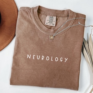 K&ouml;nnte beinhalten: Ein braunes T-Shirt mit dem Wort "NEUROLOGY" in wei&szlig; auf der Vorderseite. Das Shirt ist gefaltet und liegt flach auf einer wei&szlig;en Oberfl&auml;che. Ein brauner Hut und eine silberne Halskette sind ebenfalls auf dem Bild zu sehen.