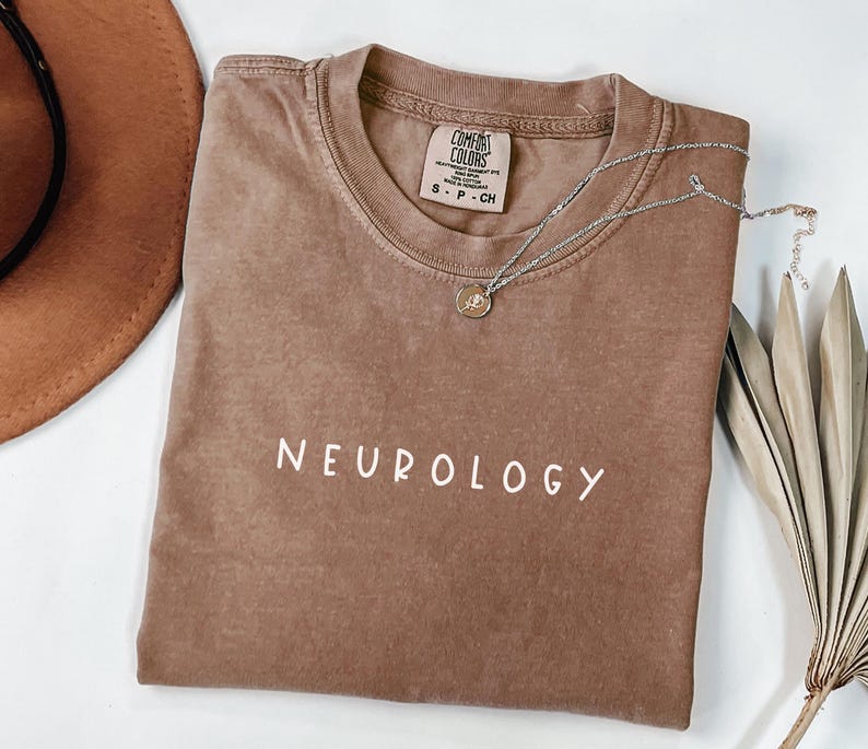 K&ouml;nnte beinhalten: Ein braunes T-Shirt mit dem Wort "NEUROLOGY" in wei&szlig; auf der Vorderseite. Das Shirt ist gefaltet und liegt flach auf einer wei&szlig;en Oberfl&auml;che. Ein brauner Hut und eine silberne Halskette sind ebenfalls auf dem Bild zu sehen.