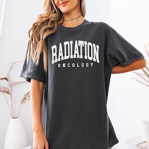 May include: Dark grey t-shirt featuring the text "RADIATION ONCOLOGY" in white, bold lettering. The shirt has a crew neck and short sleeves. The fit is loose and relaxed.