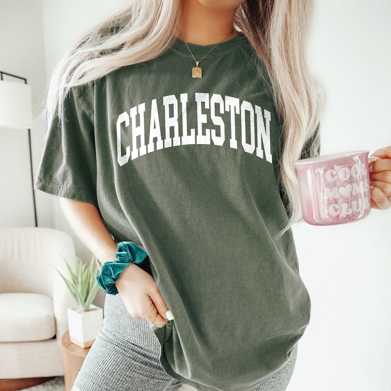 May include: A green oversized t-shirt with the word "CHARLESTON" printed in white on the front. The shirt is being worn by a person holding a pink mug with the text "Cool Moms Club" printed on it.