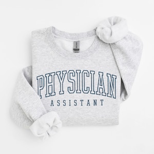 K&ouml;nnte beinhalten: Grauer Pullover mit dem Text "PHYSICIAN ASSISTANT" in dunkelblauer Schrift.