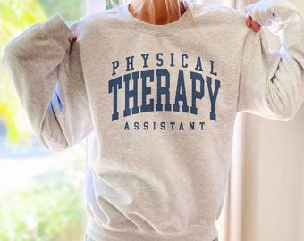 Op de afbeelding: Een lichtgrijze sweatshirt met de tekst "PHYSICAL THERAPY ASSISTANT" in marineblauwe letters. De sweatshirt heeft lange mouwen en een ronde hals.