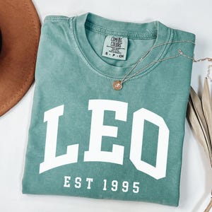 May include: A teal Comfort Colors t-shirt with the word "LEO" in large white block letters and "EST 1995" below. A gold necklace with a small pendant rests on the shirt. A brown hat and dried plant are in the background.