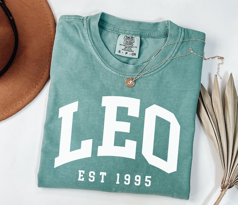 May include: A teal Comfort Colors t-shirt with the word "LEO" in large white block letters and "EST 1995" below. A gold necklace with a small pendant rests on the shirt. A brown hat and dried plant are in the background.