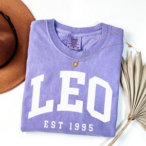 May include: A lavender Comfort Colors crew neck t-shirt with the word "LEO" in large white block letters and "EST 1995" below. A gold necklace with a pendant and a brown hat are also in the image.