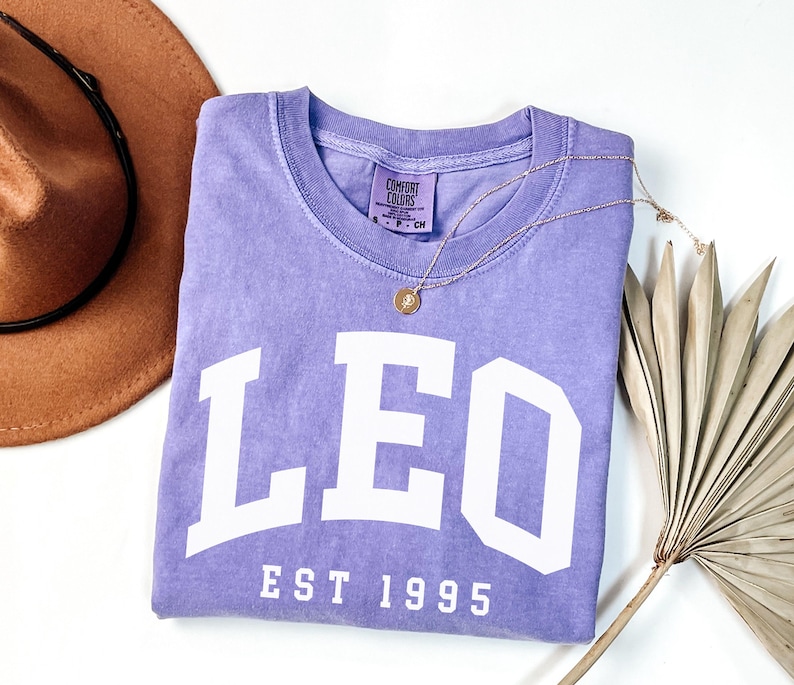 May include: A lavender Comfort Colors crew neck t-shirt with the word "LEO" in large white block letters and "EST 1995" below. A gold necklace with a pendant and a brown hat are also in the image.