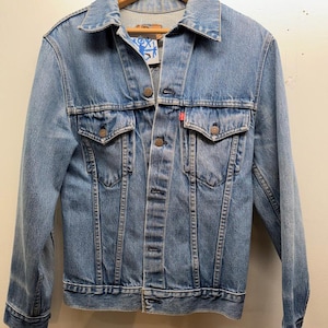 May include: A white ceramic mug with a black handle and the text "But First, Coffee" in a bold, black font. The mug is set against a blurred background, suggesting a cozy setting. The mug appears to be approximately 4 inches tall.