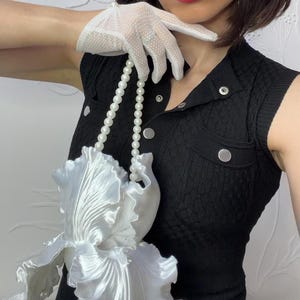 Op de afbeelding: Een witte satijnen handtas in de vorm van een grote irisbloem met een handvat van parelkralen. De tas wordt vastgehouden door een hand met een witte kanten handschoen. De outfit bevat een zwarte mouwloze top met zilveren knopen en een witte birdcage-sluier.