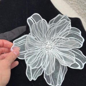 Op de afbeelding: Een grote, gelaagde witte bloemaccessoire gemaakt van doorschijnende stof met fijne witte lijndetails en een centrum van drie kleine, ronde witte parels, geplaatst tegen een achtergrond van zwarte stof.