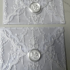 Op de afbeelding: Twee rechthoekige witte kanten enveloppen met een gedetailleerd bloemmotief en een centraal wit waszegel. Elk waszegel is voorzien van de hoofdletters MB in een cirkelvormig ontwerp, waarmee de klep van de delicate kanten envelop wordt gesloten.