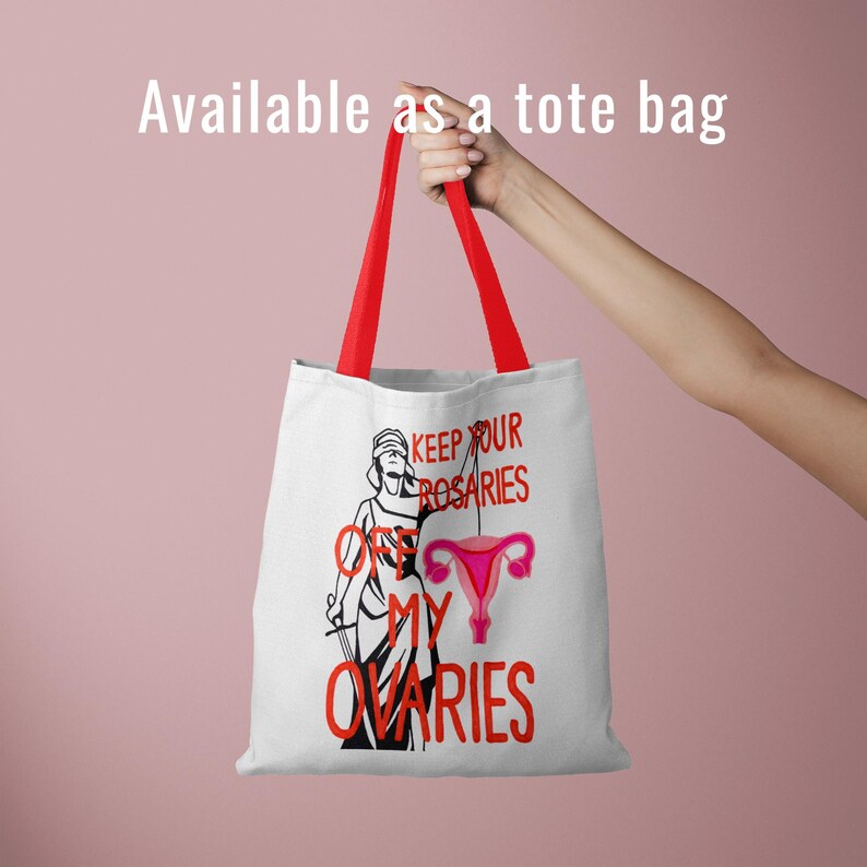 A white canvas tote bag with red handles being held against a pink background. The bag features the "Keep Your Rosaries Off My Ovaries" design. Text at the top reads: "Available as a tote bag."