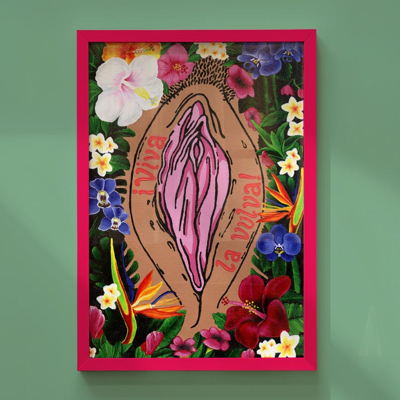 The "¡Viva la vulva!" art print with a medium-tan skin tone background and coral-red typography, displayed in a hot pink frame against a sage green wall.