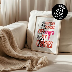 Pro-choice feminist art print in a light wood frame resting on a beige textured sofa. Sunlight filters across the scene, highlighting the red lettering and pink reproductive health illustration.