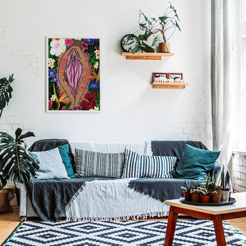 A large-scale version of the feminist vulva art print leaning against the floor in a sunlit, modern living room next to a white radiator and a large Monstera plant.