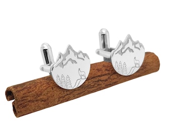 May include: A pair of silver cufflinks featuring a mountain range design with a deer and trees. The cufflinks are round and rest on a brown cinnamon stick. The design is etched into the silver.