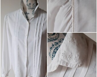 May include: A white blouse with a high, stand-up collar and long sleeves. The front of the blouse has vertical pleats, and the cuffs are decorated with embroidery. The fabric looks like a light cotton or linen.