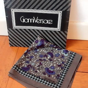 Puede incluir: Un pañuelo de seda marrón con un estampado floral en tonos de morado, azul y blanco. El pañuelo tiene un borde de pata de gallo negro y blanco y el texto "Gianni Versace" está impreso en la esquina inferior derecha.