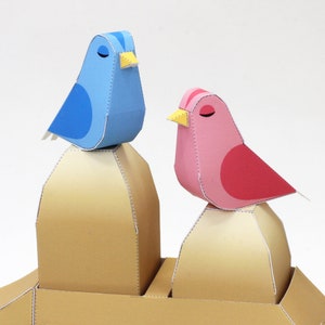May include: Two papercraft birds perched on top of two brown papercraft mounds. The bird on the left is blue with a yellow beak. The bird on the right is pink with a red belly and a yellow beak.