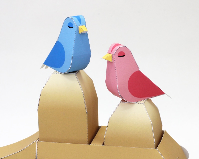 May include: Two papercraft birds perched on top of two brown papercraft mounds. The bird on the left is blue with a yellow beak. The bird on the right is pink with a red belly and a yellow beak.