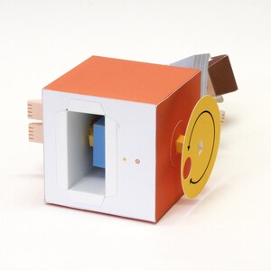 May include: A papercraft cube featuring an orange top and white sides. One side has a rectangular cutout revealing a blue block, a small yellow star, and a red letter G. A yellow circular dial with a black arrow and red dot is attached to the side.