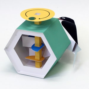 May include: A hexagonal shaped, teal and white paper toy with a yellow, blue, and orange center piece. The toy has a yellow circular top with a black arrow and a black and white bird figure attached to the side.