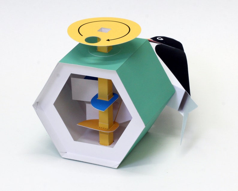 May include: A hexagonal shaped, teal and white paper toy with a yellow, blue, and orange center piece. The toy has a yellow circular top with a black arrow and a black and white bird figure attached to the side.
