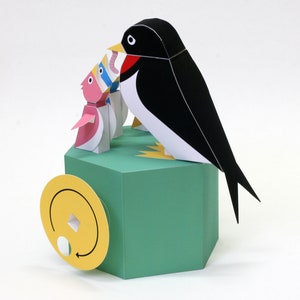 May include: A papercraft model of a swallow with a black and white body, perched on a green hexagonal base. The swallow is holding a pink and white fish in its beak. The base has a yellow circle with a white circle and an arrow on it.