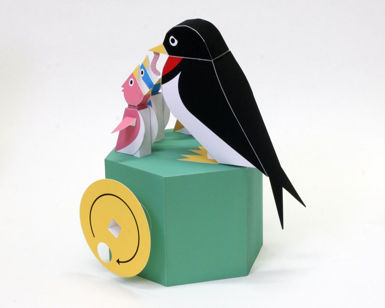 May include: A papercraft model of a swallow with a black and white body, perched on a green hexagonal base. The swallow is holding a pink and white fish in its beak. The base has a yellow circle with a white circle and an arrow on it.