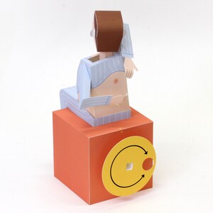 May include: A paper craft figure with brown hair wearing light blue striped pyjamas, seated on a solid orange cube. The cube features a yellow circular dial on the front with two black arrows indicating a rotational direction and a square centre hole.
