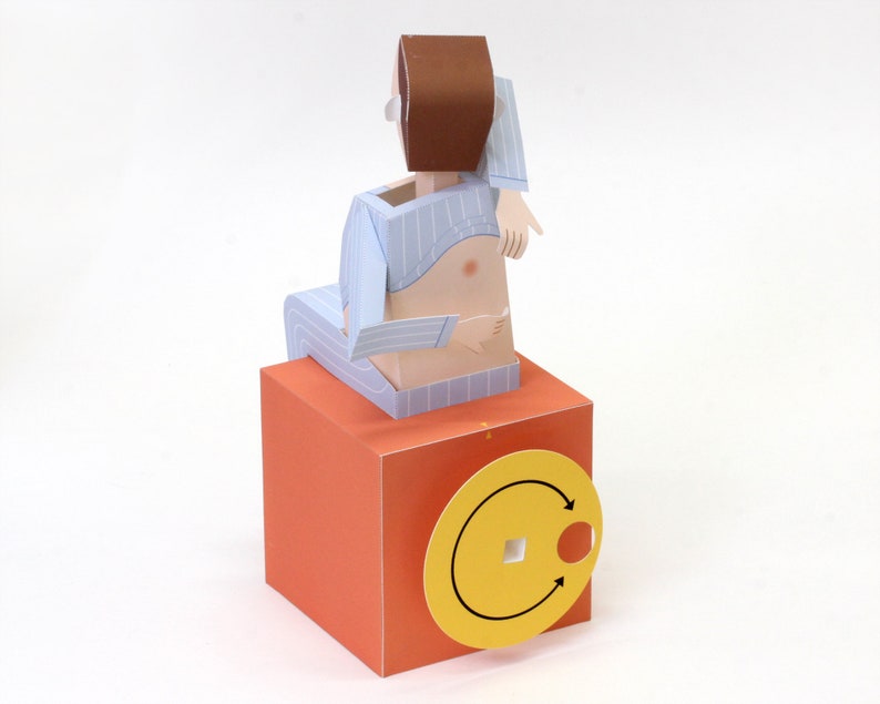 May include: A paper craft figure with brown hair wearing light blue striped pyjamas, seated on a solid orange cube. The cube features a yellow circular dial on the front with two black arrows indicating a rotational direction and a square centre hole.