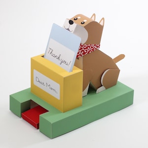 Peut inclure: Un présentoir en papier représentant un chien marron avec un bandana rouge à motifs, assis à côté d'une boîte aux lettres jaune. La boîte porte une étiquette blanche avec le texte Dear Mom, et une carte bleu clair avec le texte Thank you. Un levier rouge dépasse de la base verte.