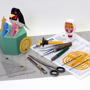 May include: A papercraft penguin model kit with instructions, tools, and glue. The kit includes a penguin with three chicks, a ruler, a compass, a cutting tool, and a glue bottle. The glue bottle is labeled "木工用" in Japanese, which means "woodworking glue".