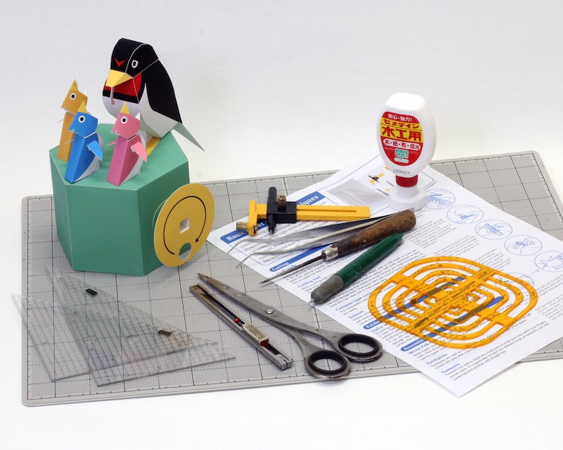 May include: A papercraft penguin model kit with instructions, tools, and glue. The kit includes a penguin with three chicks, a ruler, a compass, a cutting tool, and a glue bottle. The glue bottle is labeled "木工用" in Japanese, which means "woodworking glue".