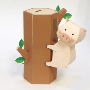 May include: A papercraft piggy bank shaped like a brown hexagonal tree trunk with a coin slot on top. A light pink pig figure with small black eyes and a snout is attached to the side of the tree, which features small green paper leaves.