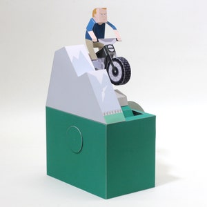 May include: A papercraft model of a person riding a motorbike over a mountain. The model is made of paper and is painted in a variety of colours, including green, white, and black.