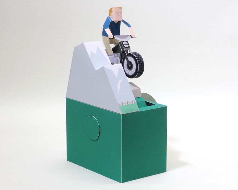 May include: A papercraft model of a person riding a motorbike over a mountain. The model is made of paper and is painted in a variety of colours, including green, white, and black.