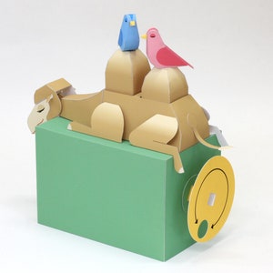 May include: A papercraft camel with a blue bird and a pink bird on its back. The camel is lying down on a green base with a yellow wheel.