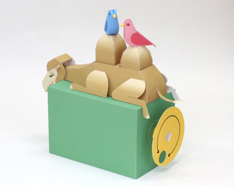 May include: A papercraft camel with a blue bird and a pink bird on its back. The camel is lying down on a green base with a yellow wheel.