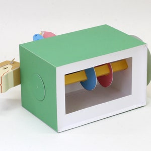 May include: A green box with a white window showing a colorful, rotating, paper toy inside. The toy is made of yellow, blue, and pink paper and is attached to a white rod. The box is open on one side, revealing the toy.