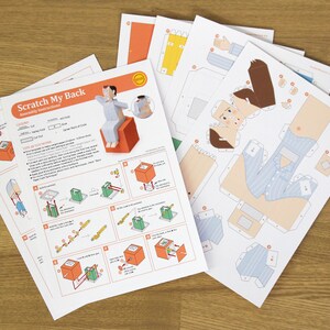 May include: A set of paper craft templates titled Scratch My Back with assembly instructions and printed parts for a character in blue striped pyjamas sitting on an orange cube. The sheets include diagrams, cut lines, and fold markers.